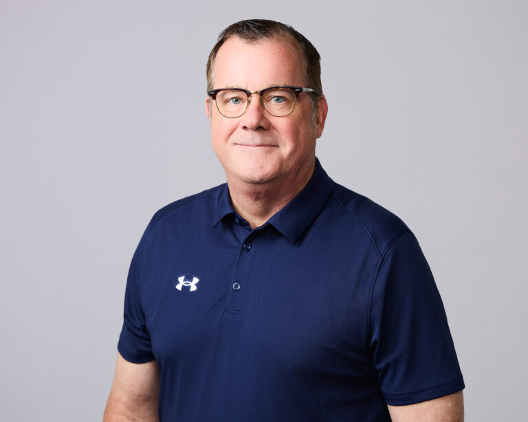 Man wearing glasses and a navy blue Under Armour polo shirt standing against a plain light gray background.