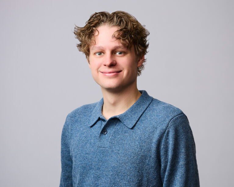 A young man with curly hair wearing a blue collared sweater stands in front of a plain light gray background, smiling at the camera.