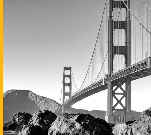 Black-and-white photo of the Golden Gate Bridge spanning over water, with rocky shoreline and hills visible in the background.