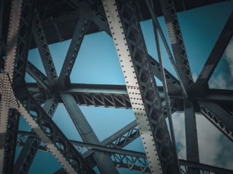Close-up view of intersecting steel beams and rivets forming the structure of a bridge, with blue sky and clouds visible in the background.