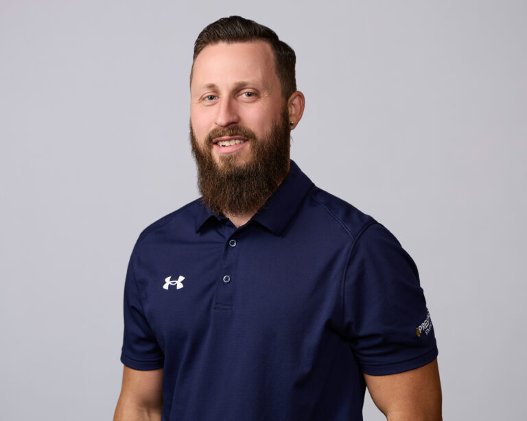 A man with a beard and short hair wearing a navy blue Under Armour polo shirt stands against a plain light gray background.