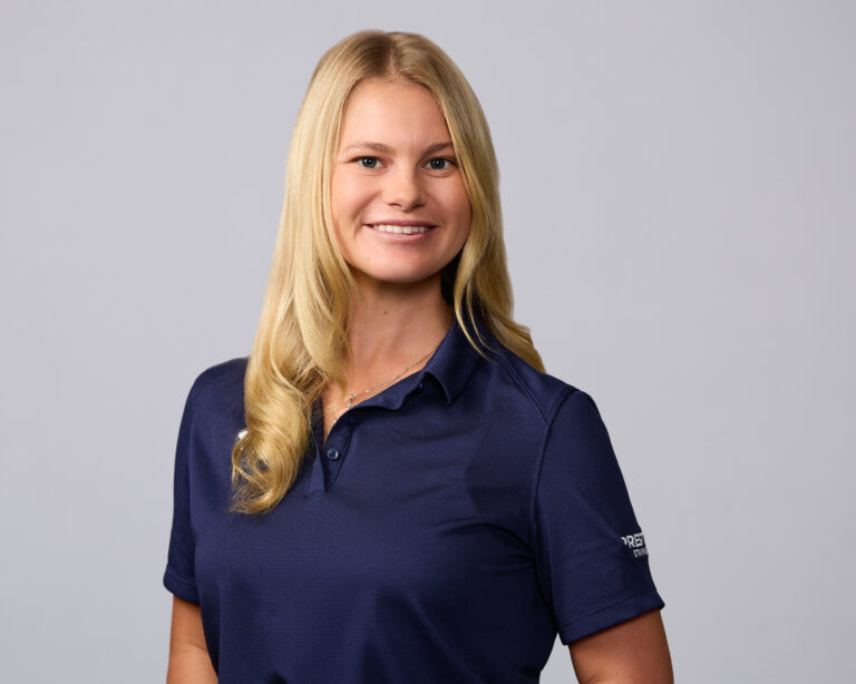A woman with long blonde hair wearing a navy blue collared shirt stands against a plain light gray background, smiling at the camera.
