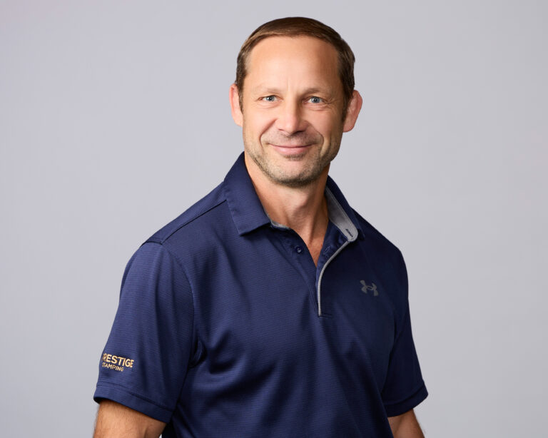 Man with short brown hair wearing a navy blue Under Armour polo shirt stands smiling against a plain light gray background.