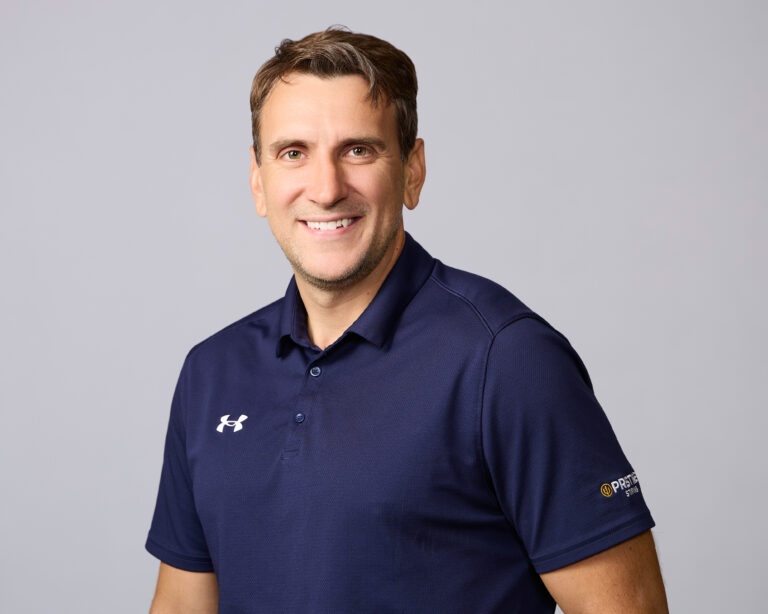 Smiling man wearing a navy blue polo shirt with the Under Armour logo and a Premier logo on the sleeve, standing against a plain light gray background.