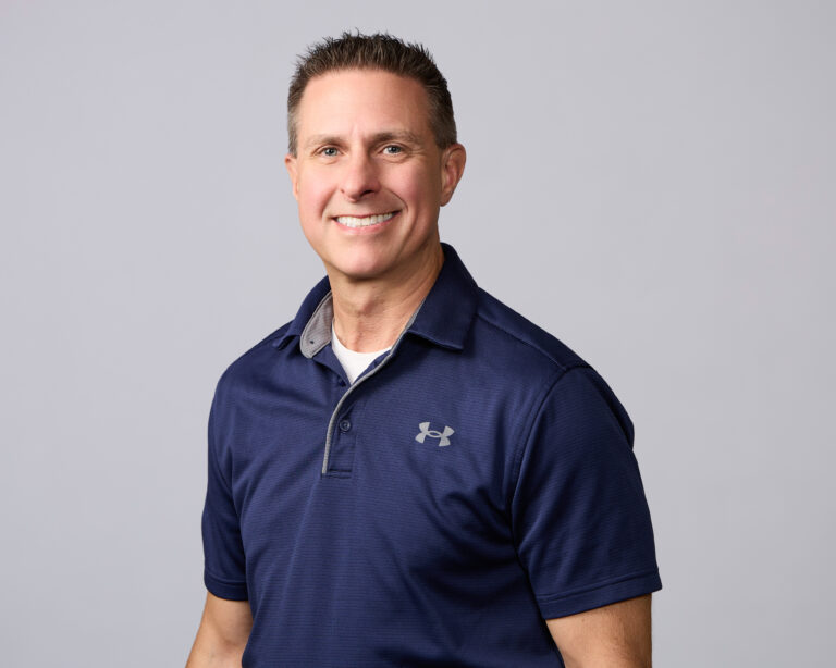 Smiling man wearing a dark blue collared Under Armour shirt standing in front of a plain light gray background.