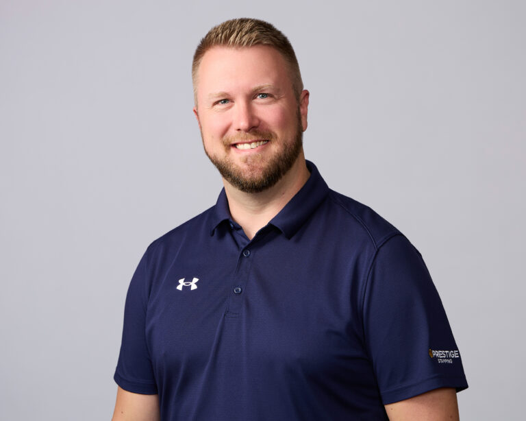 A man with short blond hair and a beard smiles, wearing a navy polo shirt with Under Armour and Prestige Staffing logos, against a plain light background.