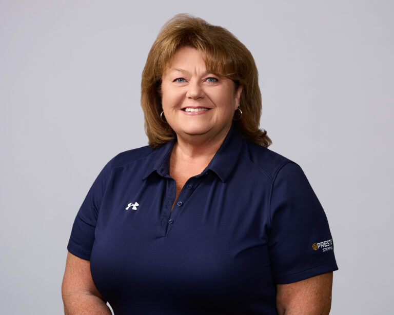 A woman with shoulder-length light brown hair wearing a navy blue polo shirt stands in front of a plain, light grey background, smiling at the camera.