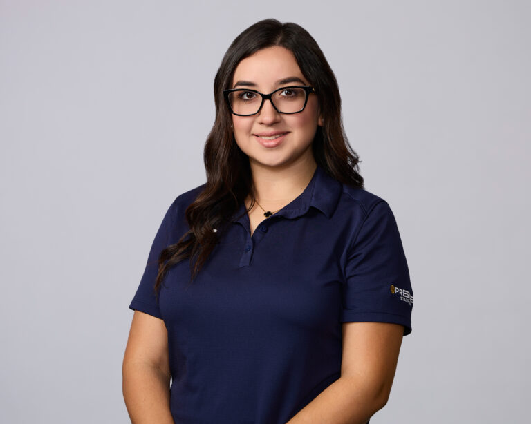 A woman with long dark hair and glasses wearing a navy blue polo shirt stands against a plain light background, facing the camera and smiling.