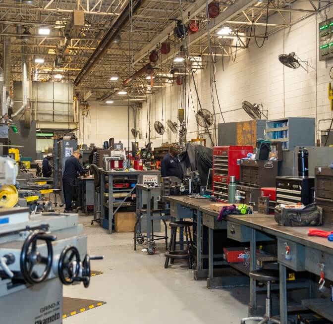 A large, well-lit industrial workshop with multiple workbenches, toolboxes, machinery, and workers engaged in various tasks.