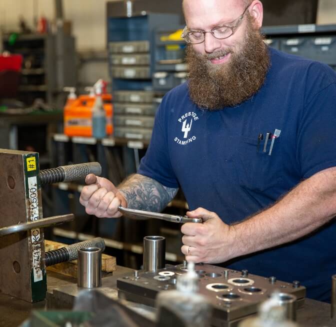 A man wearing glasses and a blue shirt works with metal tools and parts in an industrial workshop setting.