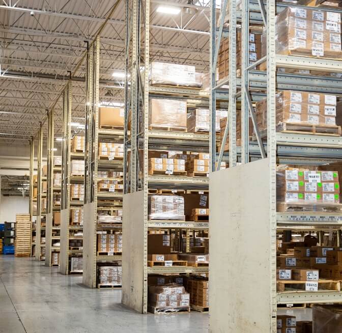 Warehouse interior with tall metal racks filled with stacked cardboard boxes and pallets, under bright overhead lighting.