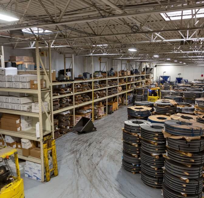Industrial warehouse interior with shelves of boxes on the left and tall stacks of coiled metal strips on the right; overhead lighting and high ceiling visible.