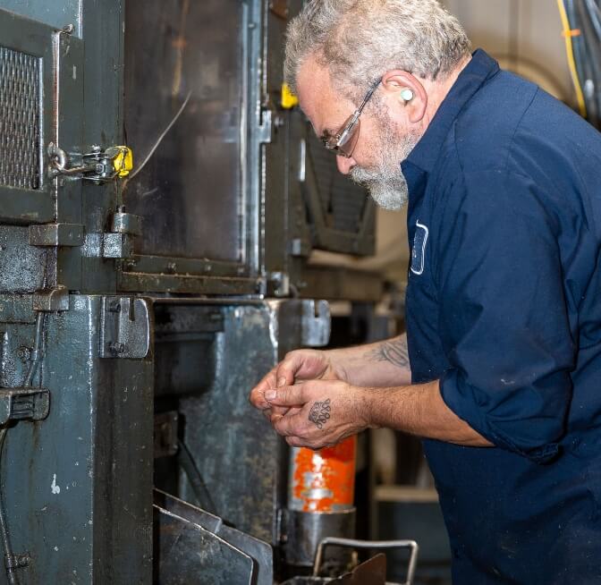 A man wearing safety glasses and earplugs operates machinery in an industrial setting, inspecting a small component in his hands.