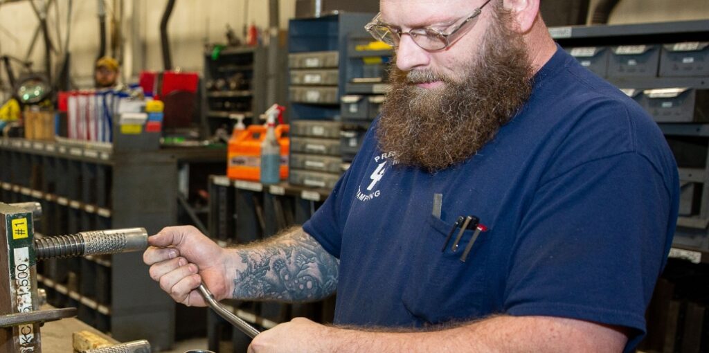 A bearded man wearing glasses works with a metal tool in a workshop, surrounded by shelves and equipment.