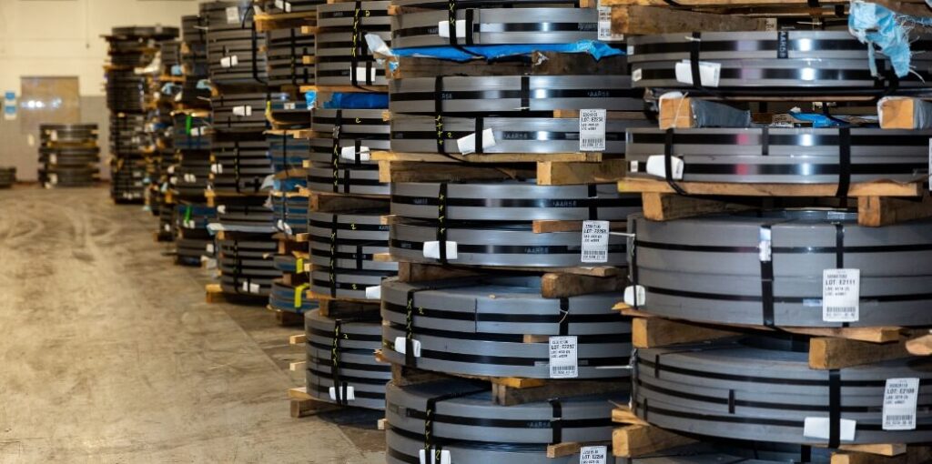 Stacks of metal coils are arranged on wooden pallets in a warehouse, with labels and packaging materials visible on the coils.
