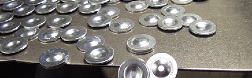 Metal discs with raised centers are scattered across a flat metallic surface in an industrial setting.