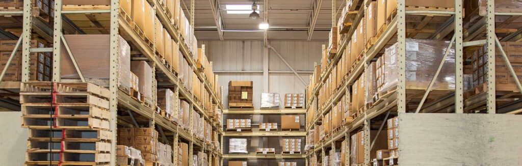 Tall shelves filled with stacked cardboard boxes and pallets line both sides of an aisle inside a large warehouse.