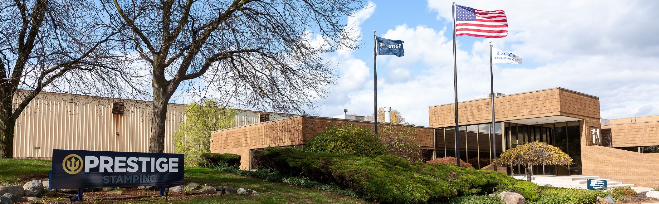 A commercial building with large windows, three flagpoles, and a sign that reads "Prestige Stamping" stands amid trees and bushes under a partly cloudy sky.