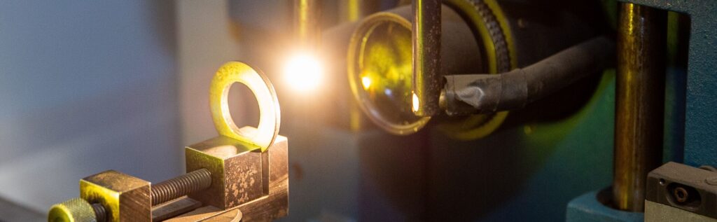 A close-up of a laser welding machine in operation, focusing on a metal ring held in place by a clamp as the laser beam is applied.