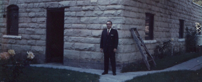 A man in a dark suit stands on a walkway outside a stone building with a ladder leaning against the wall.