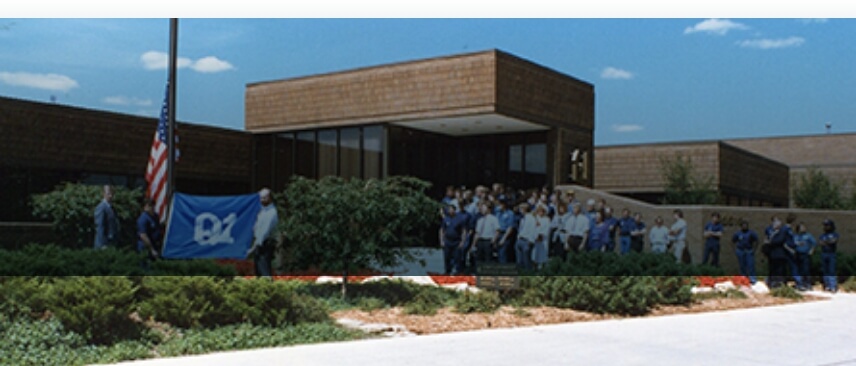 A group of people pose outside a brick office building, next to a flagpole with the U.S. flag and a blue flag with "B1" written on it.