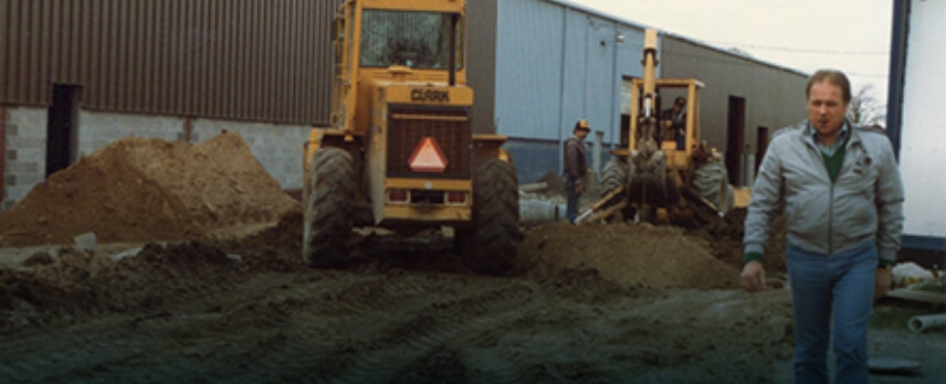 A man walks near construction machinery and dirt piles outside a large industrial building, with workers operating equipment in the background.