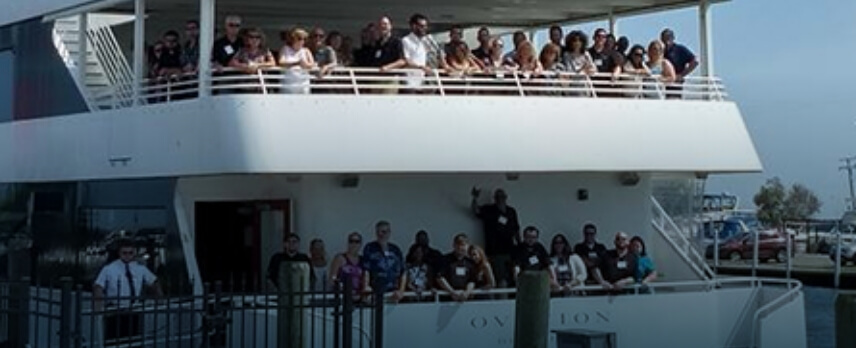 A large group of people stands on the upper and lower decks of a docked white boat, some facing the camera, with a few crew members visible.