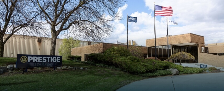 A commercial building with a “Prestige Stamping” sign in front, three flagpoles, and trees surrounding the entrance on a cloudy day.