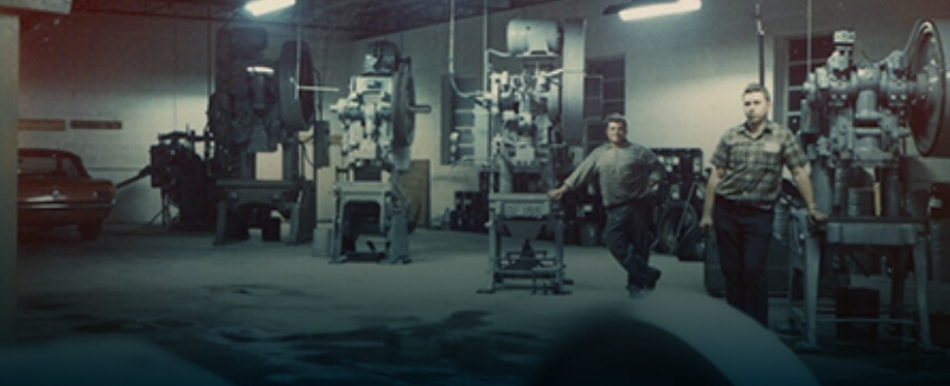 Two men walk through a dimly lit industrial workshop with several large metal machines and a parked car visible in the background.