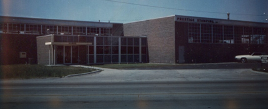 A low, rectangular industrial building with large windows labeled "PRESTIGE STAMPING" sits next to a small parking lot with one white car.