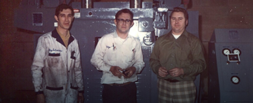 Three men stand in front of industrial machinery; two wear work jackets, one in a green shirt and plaid pants. The setting appears to be a workshop or factory.