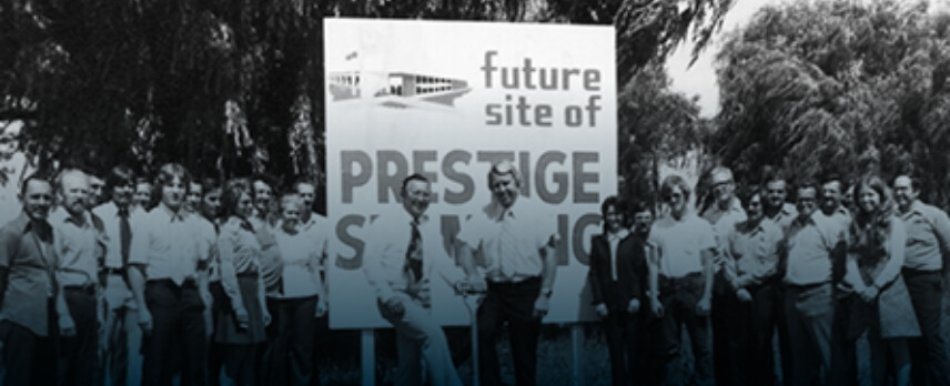 A group of people stand in front of a sign that reads "future site of Prestige Savings," with trees in the background.