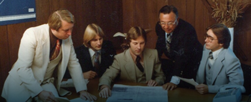 Five men in suits are gathered around a table, examining documents and discussing plans in a wood-paneled office.