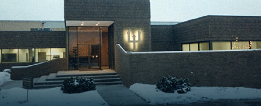 A modern brick building with large windows and a glass entrance, surrounded by snow, with a trident symbol illuminated on the exterior wall.